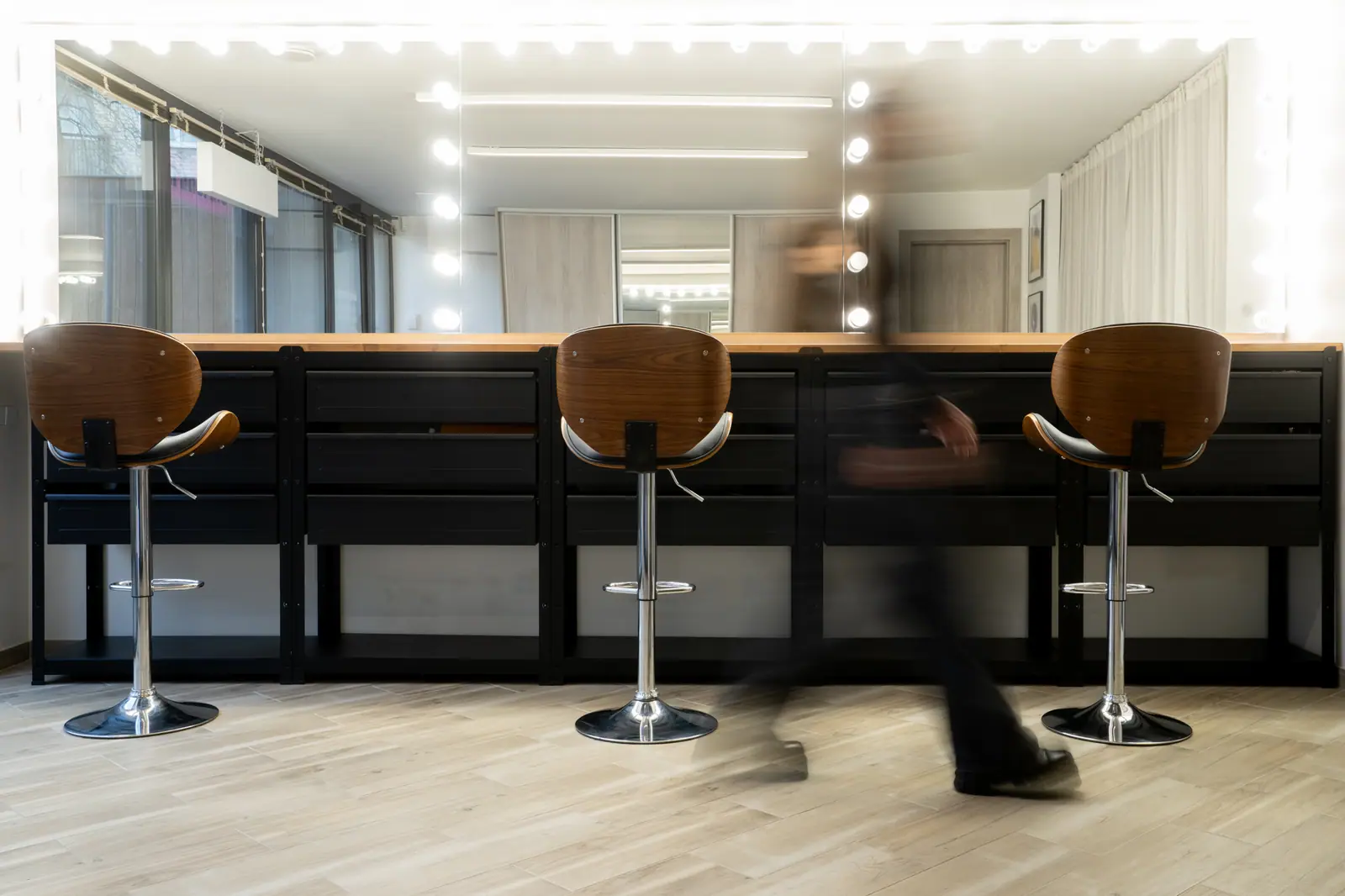 Close-up of Grimerinė's long makeup bar with a mirror framed by round vanity lights, three bar stools, and a glimpse of the street through the side window.